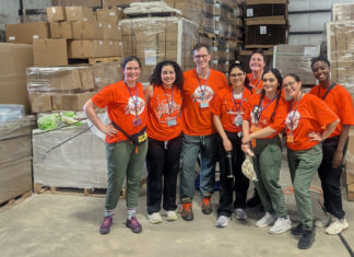 Mercer family therapy students provide behavioral healthcare to farmworkers MUSM team in matching orange shirts stand and smile in a warehouse with boxes and pallets in the background.