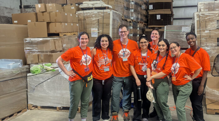 Mercer family therapy students provide behavioral healthcare to farmworkers MUSM team in matching orange shirts stand and smile in a warehouse with boxes and pallets in the background.