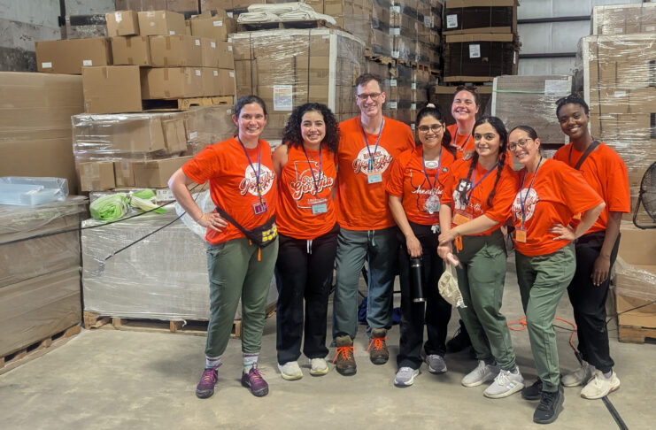 MUSM team in matching orange shirts stand and smile in a warehouse with boxes and pallets in the background.