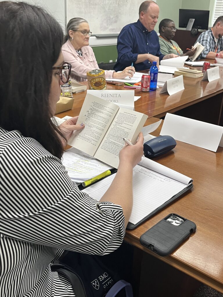 A group of adults sits at a table with books and notebooks, participating in a meeting or class discussion.