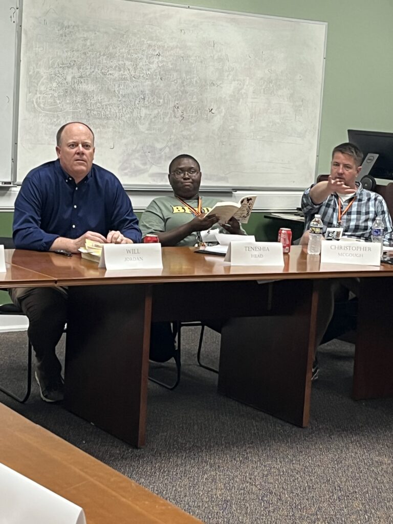 Three people sit at a conference table in a classroom, with name cards reading Will, Teneshia, and Christopher.