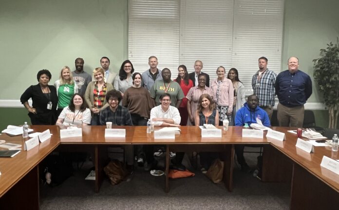 A group of adults pose for a photo in a conference room, seated and standing around wooden tables with nameplates.