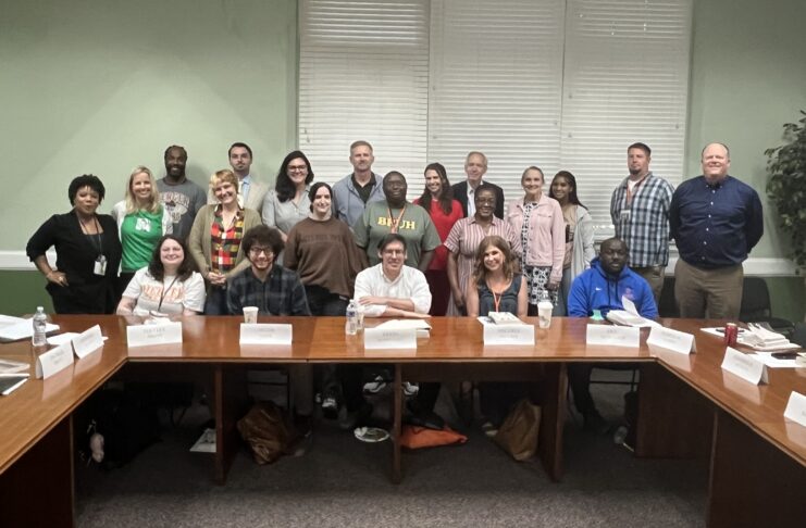 A group of adults pose for a photo in a conference room, seated and standing around wooden tables with nameplates.