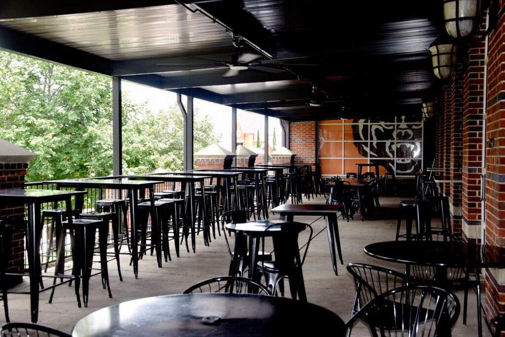 Covered outdoor patio with metal tables and chairs, brick walls, and trees visible in the background.