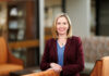 School of Medicine alumna continues tradition of mentorship as associate dean for student affairs Woman in a maroon blazer and blue shirt smiles while standing in a modern, warmly lit office lounge area.