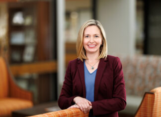 School of Medicine alumna continues tradition of mentorship as associate dean for student affairs Woman in a maroon blazer and blue shirt smiles while standing in a modern, warmly lit office lounge area.