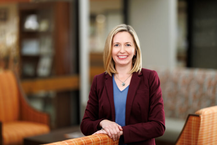 Woman in a maroon blazer and blue shirt smiles while standing in a modern, warmly lit office lounge area.