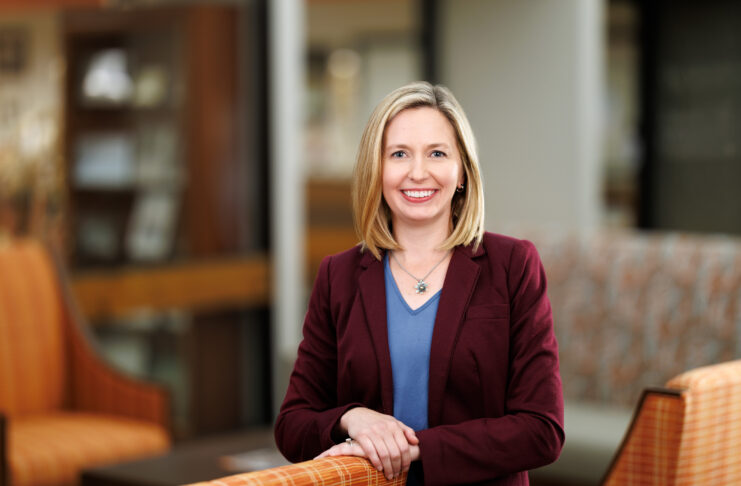 Woman in a maroon blazer and blue shirt smiles while standing in a modern, warmly lit office lounge area.