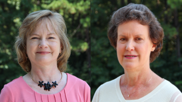 Two women stand outdoors in front of greenery, both facing the camera and smiling slightly.