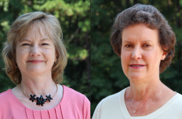 Two women stand outdoors in front of greenery, both facing the camera and smiling slightly.