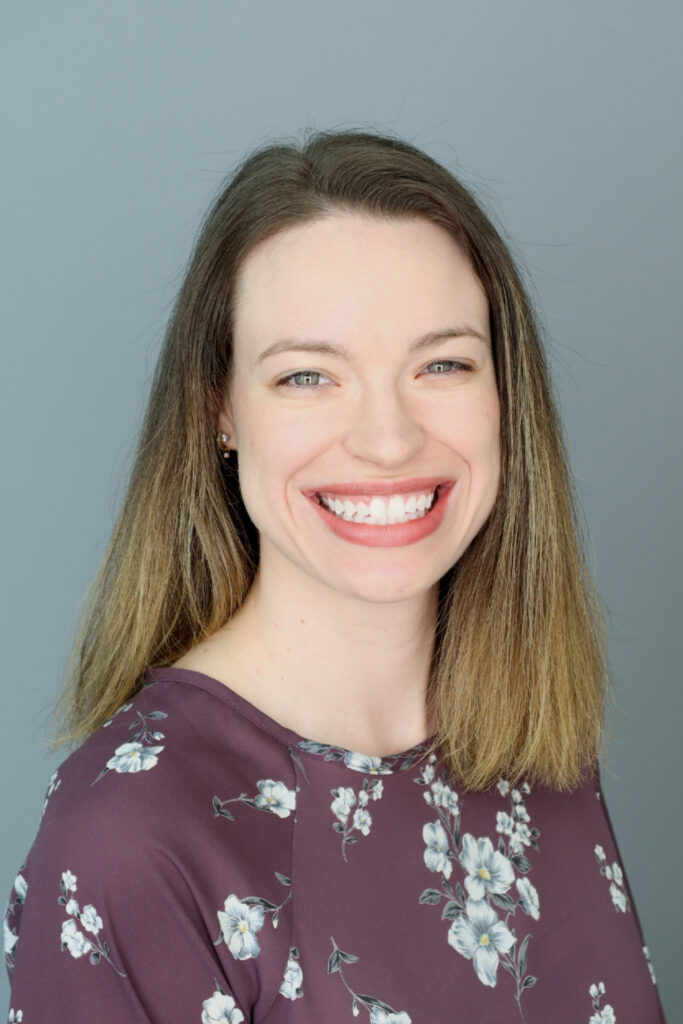 Woman with straight, shoulder-length hair wearing a purple floral top, smiling in front of a plain gray background.