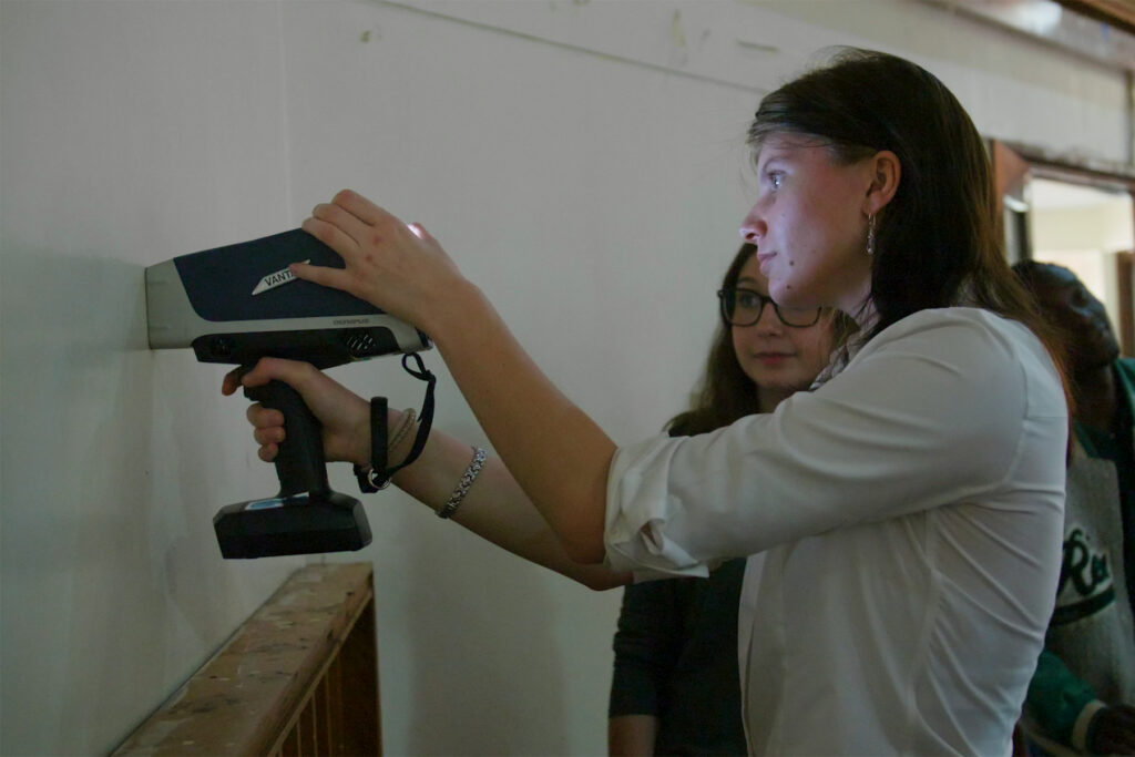 A woman uses a handheld scanning device on a white wall while another person observes beside her.