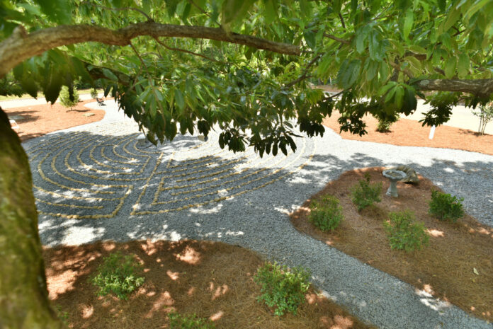 A stone labyrinth is visible beneath tree branches, surrounded by shrubs and pine straw in a garden setting.