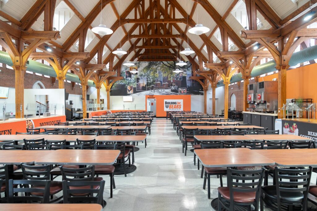 Spacious dining hall with rows of empty tables, wooden beams, orange accents, and food stations along the walls.