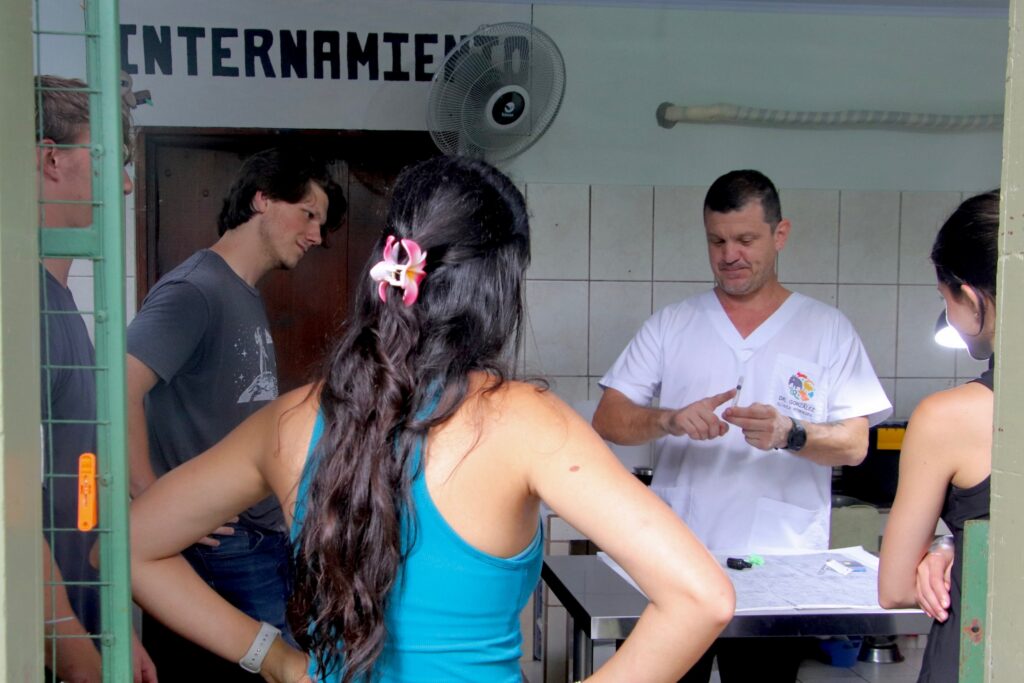 A man in a white uniform explains something to four people standing around a table in a tiled room labeled INTERNAMIENTO.