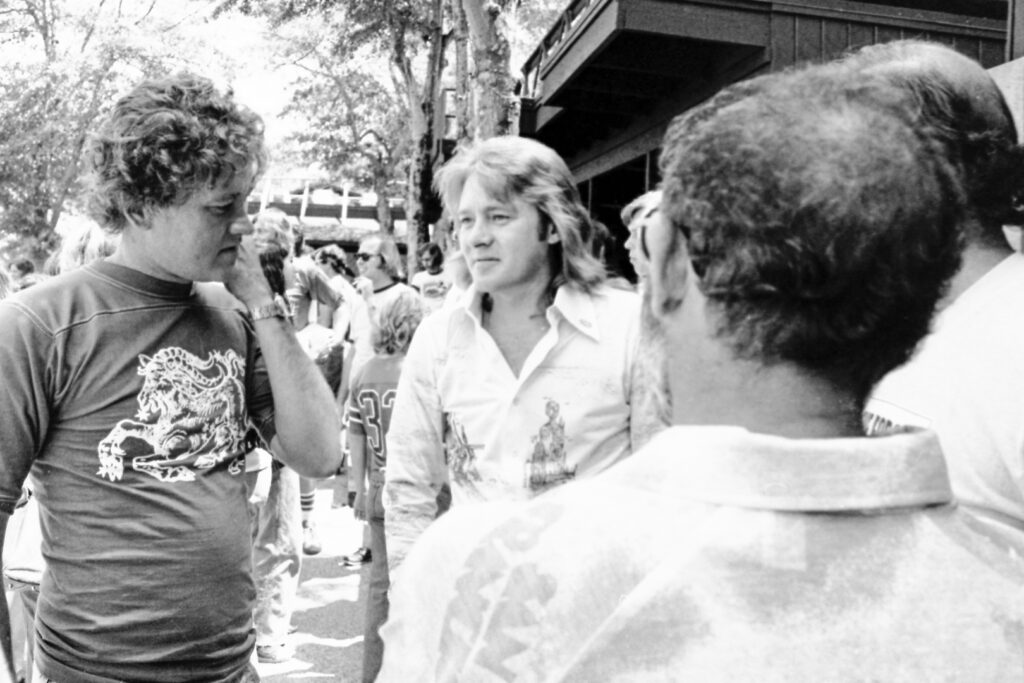 A group of men stand and talk outside in daylight, surrounded by other people and trees.