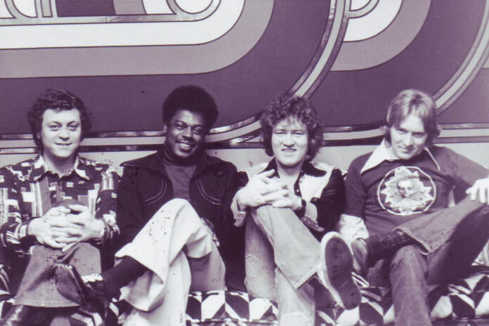 Four men sitting on a patterned couch in front of a geometric backdrop, all facing the camera and smiling.