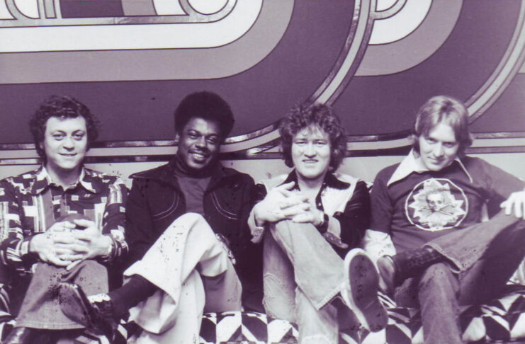 Four men sitting on a patterned couch in front of a geometric backdrop, all facing the camera and smiling.