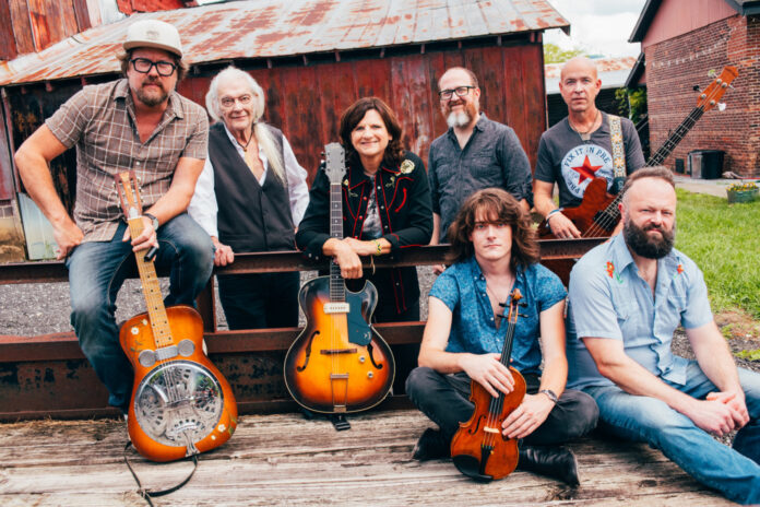 Seven people with musical instruments pose outdoors in front of a rustic barn and metal fence.