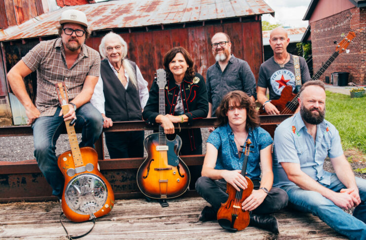 Seven people with musical instruments pose outdoors in front of a rustic barn and metal fence.