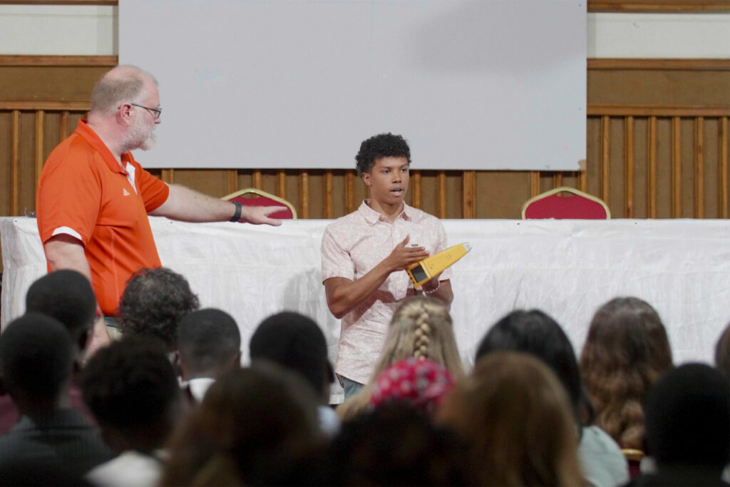 A young man holding a yellow object speaks to an audience while an older man in an orange shirt gestures beside him.