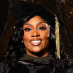 Woman wearing a black graduation cap and gown, smiling in front of a dark background.