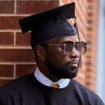 Man in graduation cap and gown with sunglasses leans against a brick wall, looking to the side.