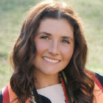 Young woman in graduation attire smiling outdoors, wearing an orange sash and honor cords around her neck.