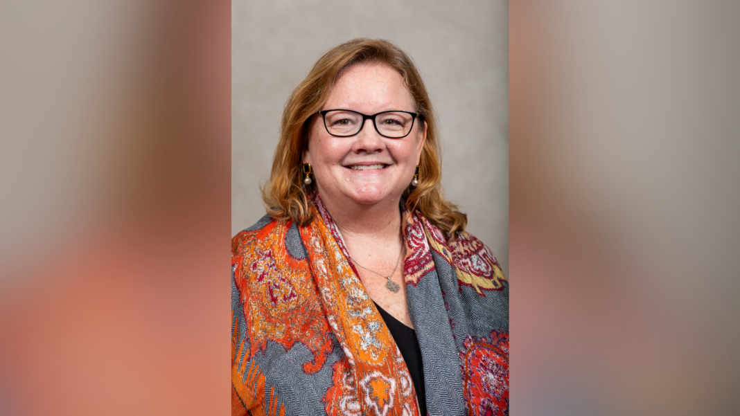 Woman with glasses and long hair, smiling, wearing an orange patterned scarf over a dark top, in front of a neutral background.