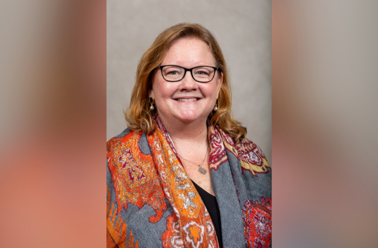 Woman with glasses and long hair, smiling, wearing an orange patterned scarf over a dark top, in front of a neutral background.