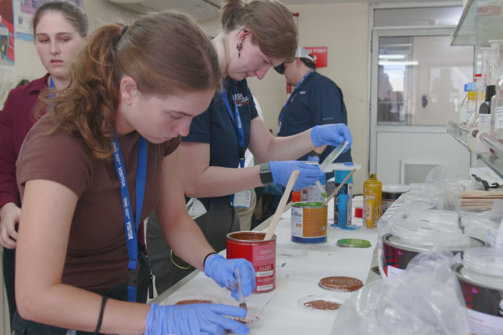 Two people wearing gloves work with paint and wood samples on a table in a laboratory setting.