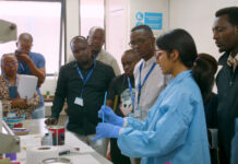 Mercer On Mission program in Kenya teaches low-cost method to test for lead in paint A group of people observe a scientist in a lab coat and gloves demonstrating an experiment in a laboratory.