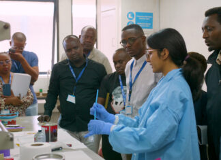 Mercer On Mission program in Kenya teaches low-cost method to test for lead in paint A group of people observe a scientist in a lab coat and gloves demonstrating an experiment in a laboratory.