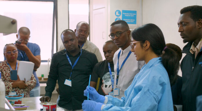 Mercer On Mission program in Kenya teaches low-cost method to test for lead in paint A group of people observe a scientist in a lab coat and gloves demonstrating an experiment in a laboratory.