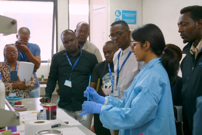 mom kenya 2026 A group of people observe a scientist in a lab coat and gloves demonstrating an experiment in a laboratory.