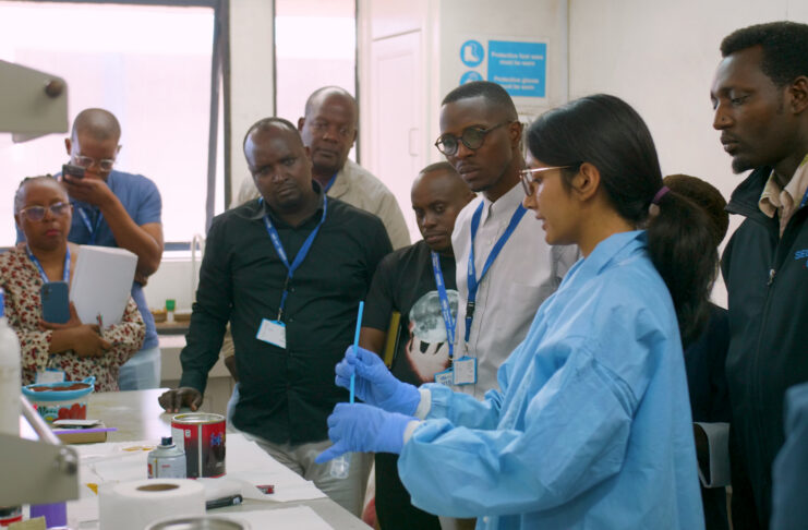 A group of people observe a scientist in a lab coat and gloves demonstrating an experiment in a laboratory.