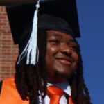 A graduate wearing a cap, tassel, and orange stole smiles outdoors in sunlight against a blue sky.