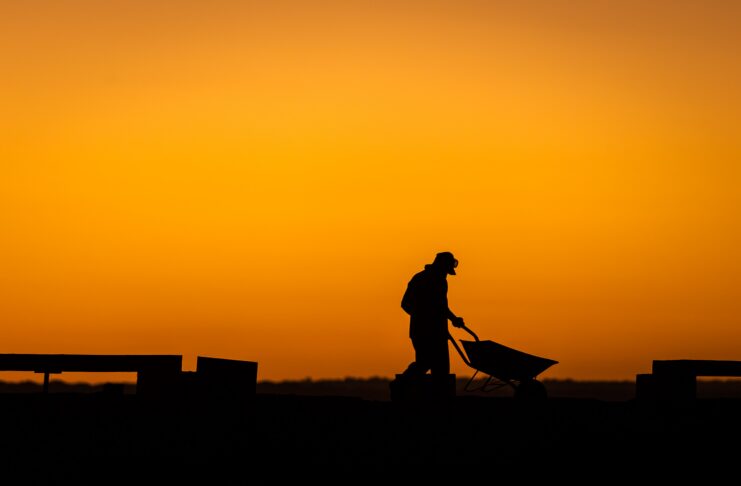 Silhouette of a person pushing a wheelbarrow at sunset, with an orange sky in the background.