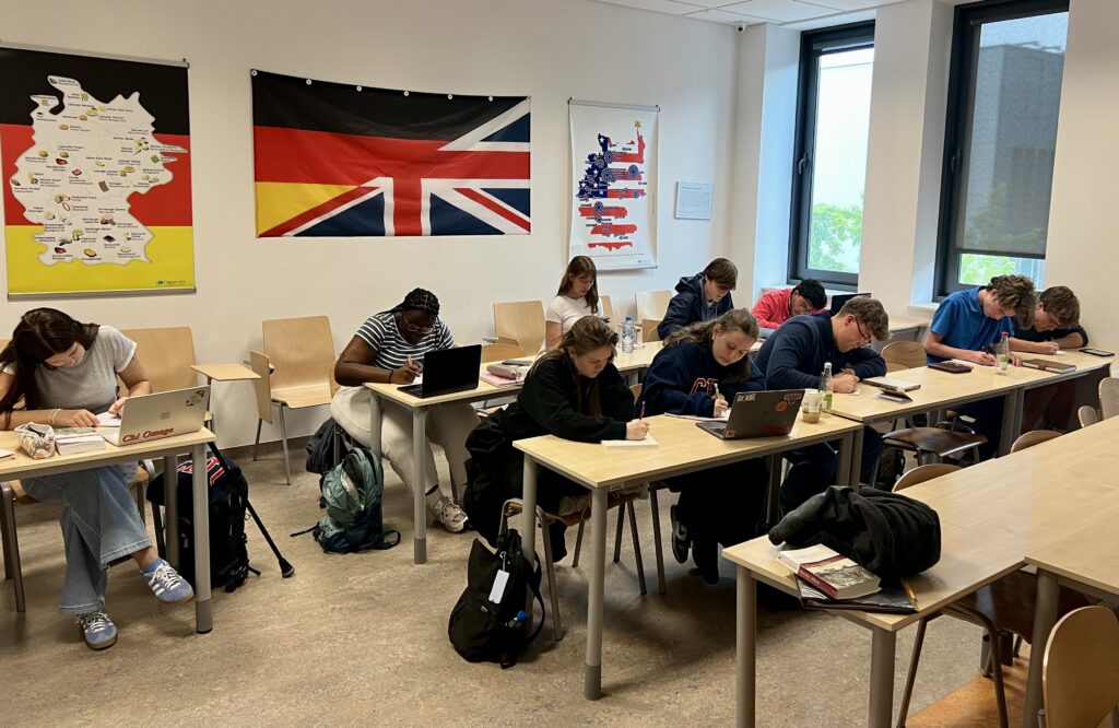 Students sitting at desks in a classroom, using laptops and notebooks, with German and UK flags on the wall.