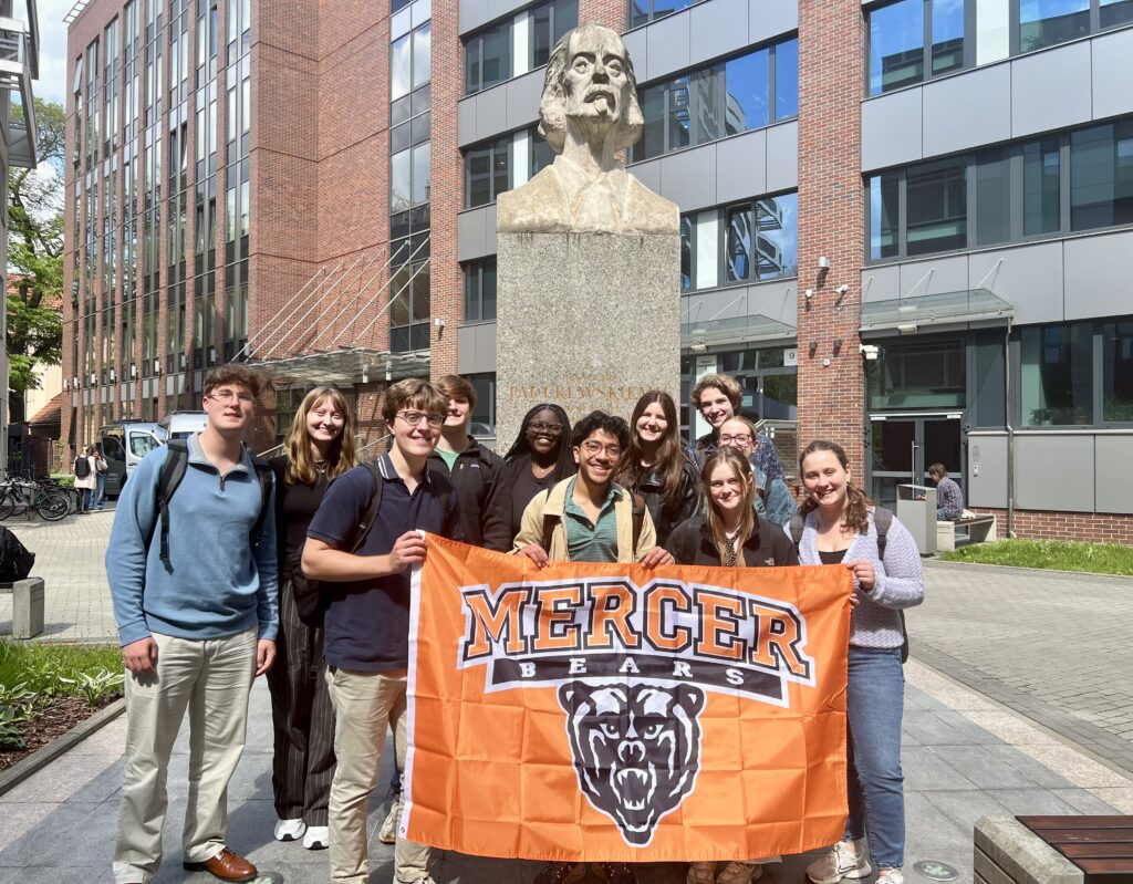 A group of students holds a Mercer Bears banner in front of a statue outside a modern academic building.