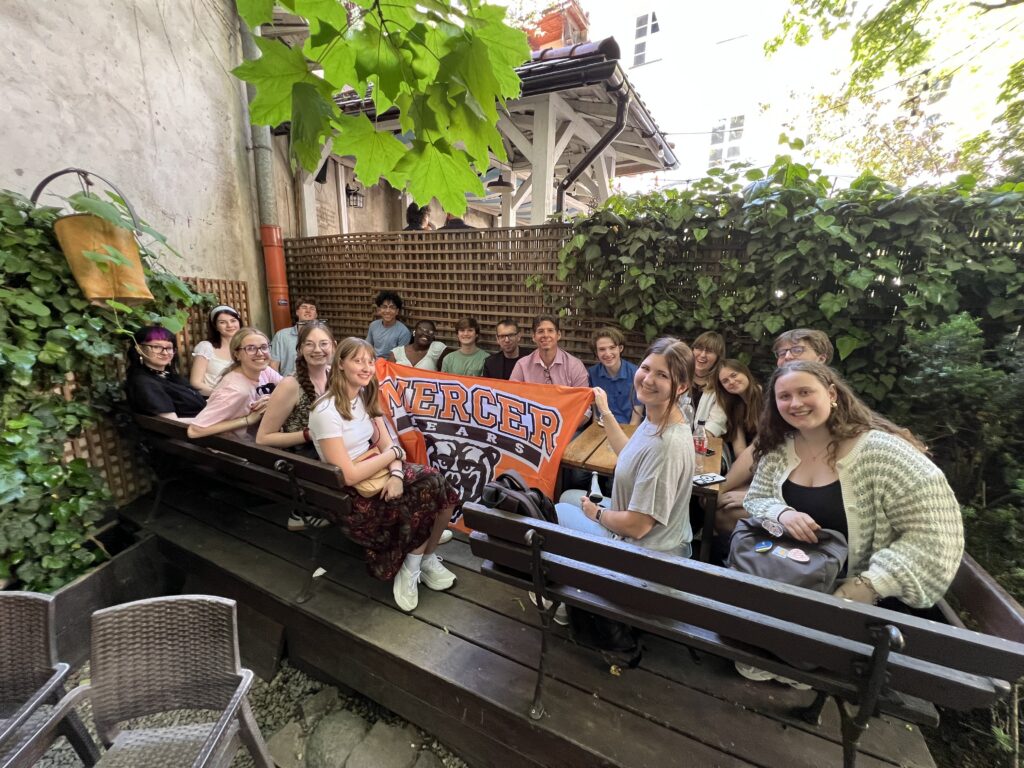A group of people sit outdoors at wooden tables holding a Mercer University banner surrounded by greenery.