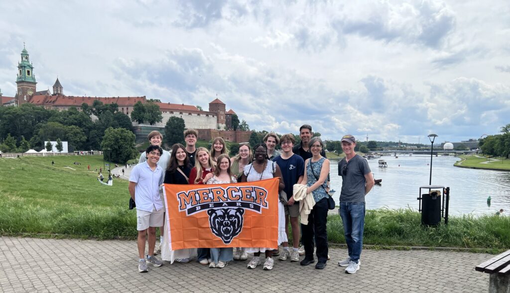 Group of people holding a Mercer University banner by a river, with historic buildings and greenery in the background.