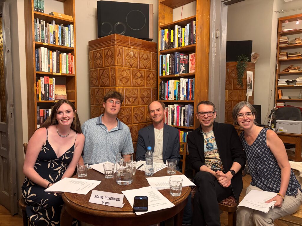 Five people sit at a round table with papers and water glasses, surrounded by bookshelves in a cozy room.