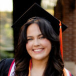 A person wearing a graduation cap and gown smiles at the camera outdoors.