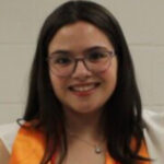 Young woman with long dark hair and glasses, wearing an orange stole and white outfit, smiling at the camera.