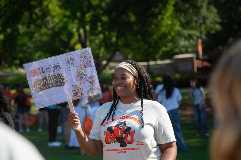 Woman smiling outdoors, holding a handmade Guardians of the Galaxy sign at a campus event with people in the background.