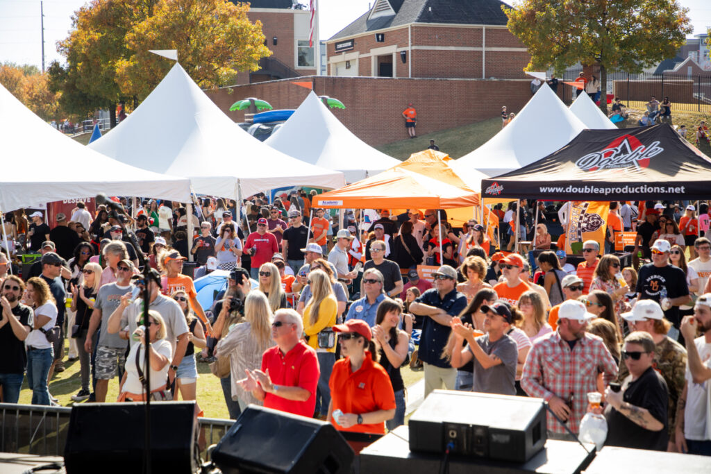 A large crowd stands outdoors at a festival with tents, stages, and people enjoying the sunny weather.