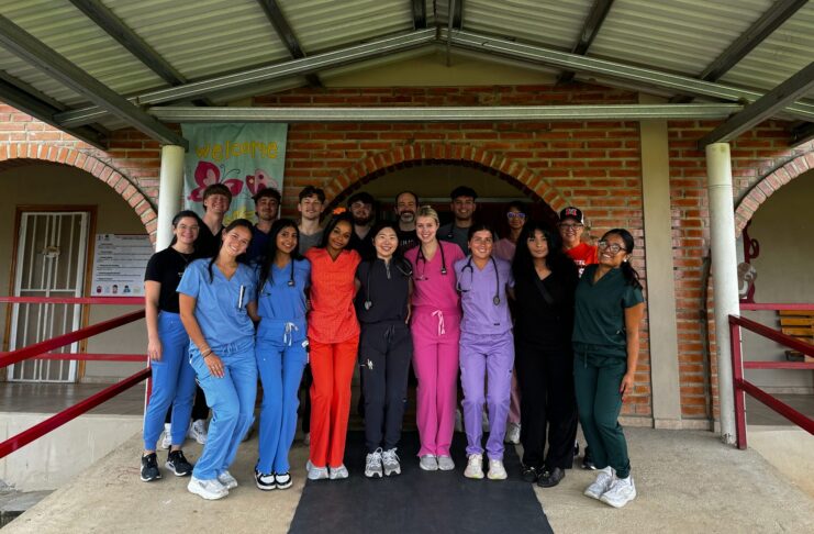 A group of people in colorful scrubs stands together under a covered walkway in front of a brick building.