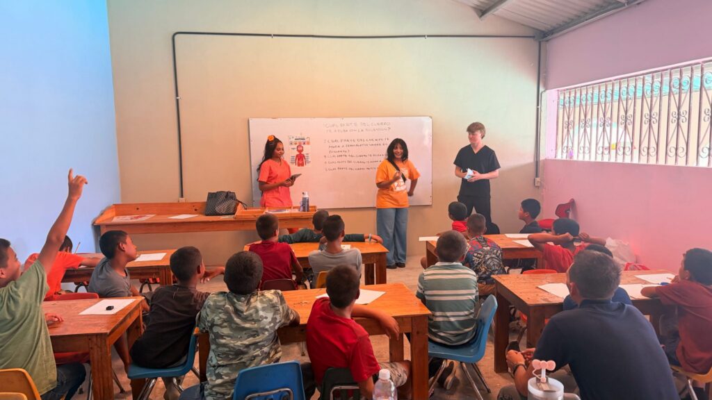 Three people stand at the front of a classroom, speaking to children who are seated at desks and raising their hands.