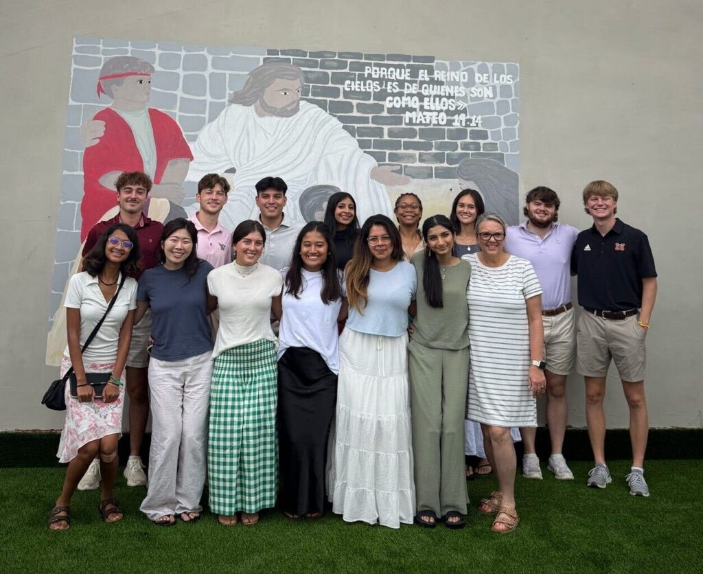A group of people stands on grass in front of a mural on a gray wall, posing and smiling for a group photo.